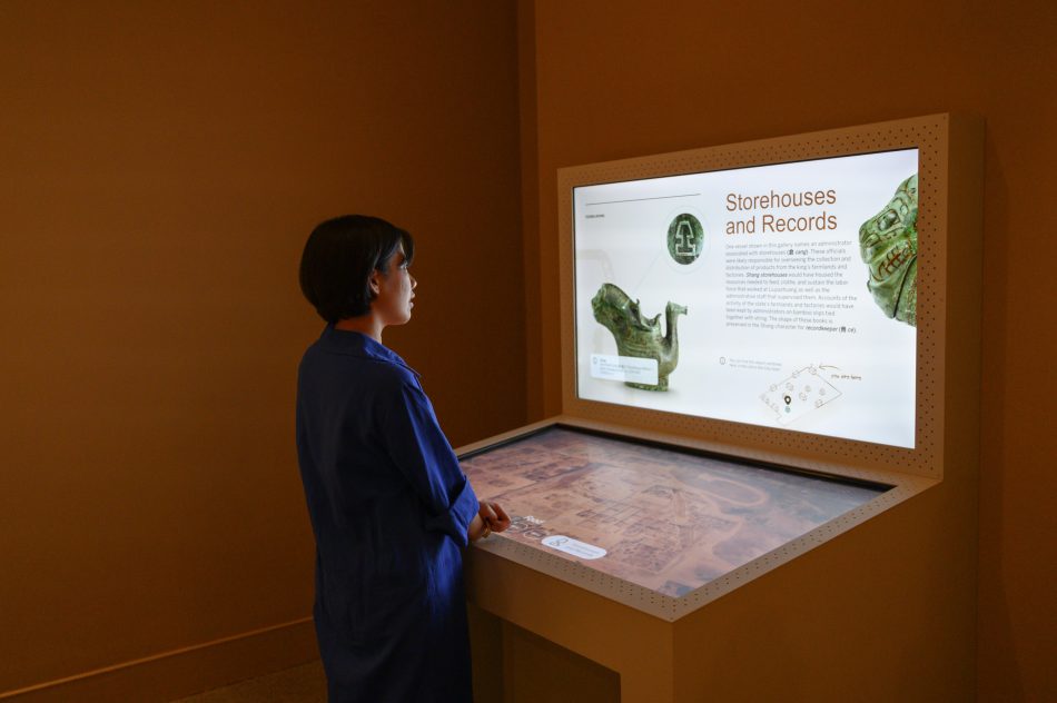 Woman standing in front of a digital kiosk with a table top touch screen and a wall mounted display.